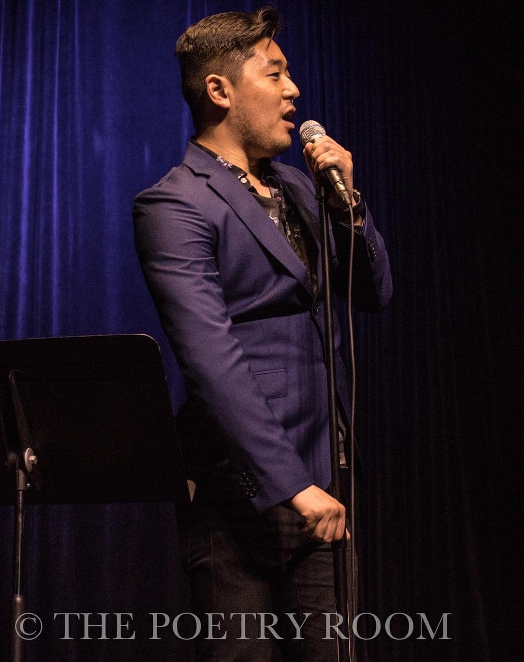 Side view of man in blue suit with old style mic in front of blue stage curtain.